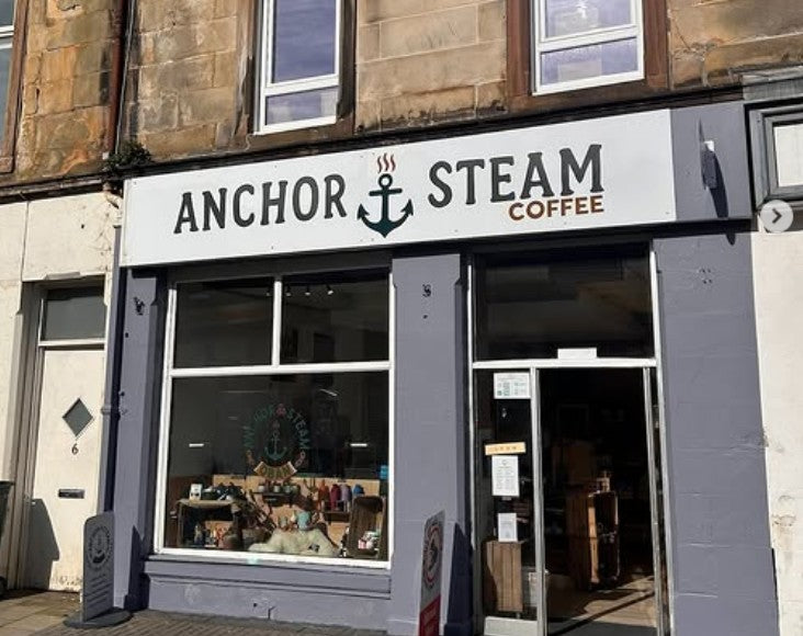 The storefront of Anchor Steam Coffee, featuring a white sign with black text and an anchor logo above a grey facade with large windows and an open door.