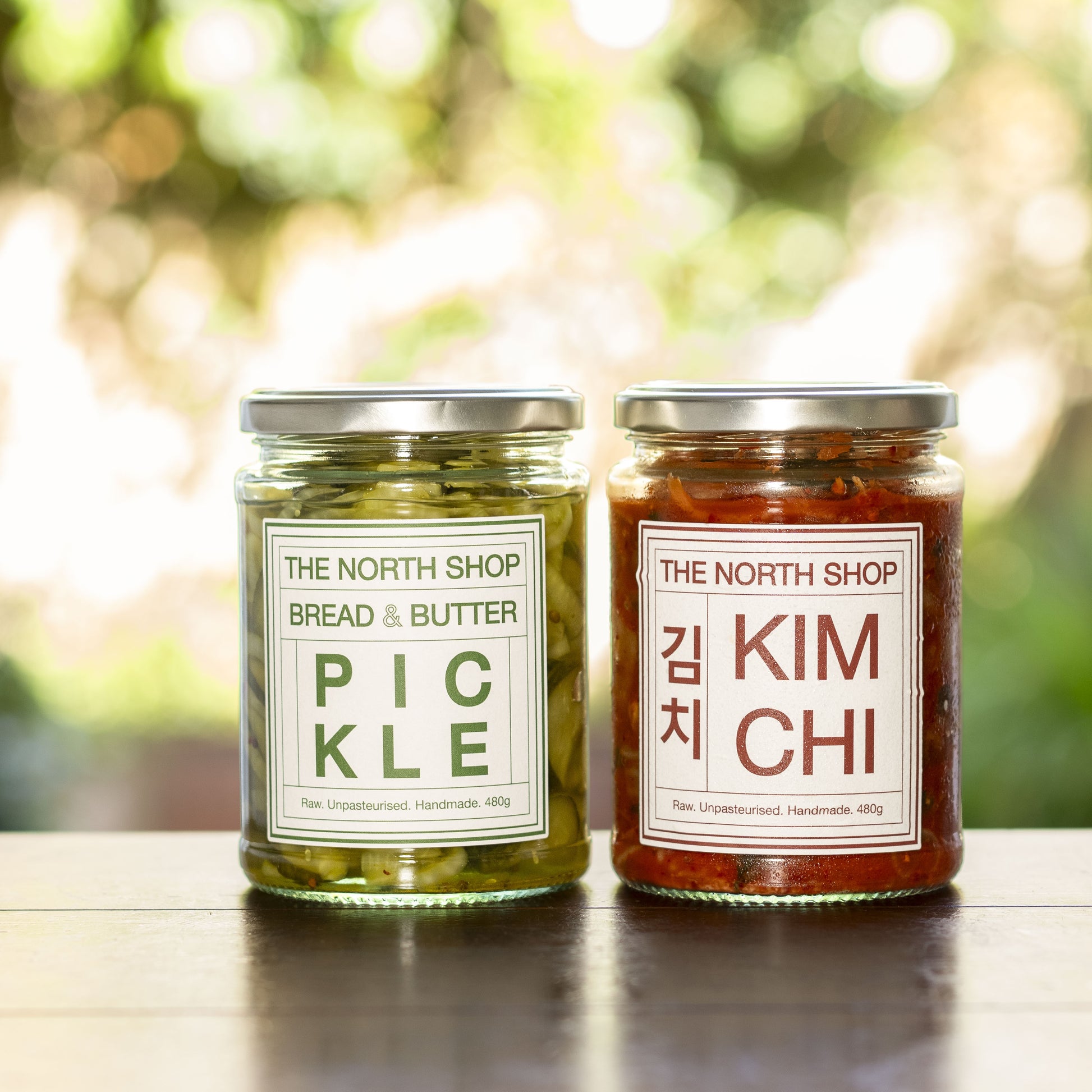 Two glass jars of The North Shop spreads on a dark wooden surface with a blurred green outdoor bokeh background. The left jar contains pickle with a green label, and the right jar contains kimchi with a red label.
