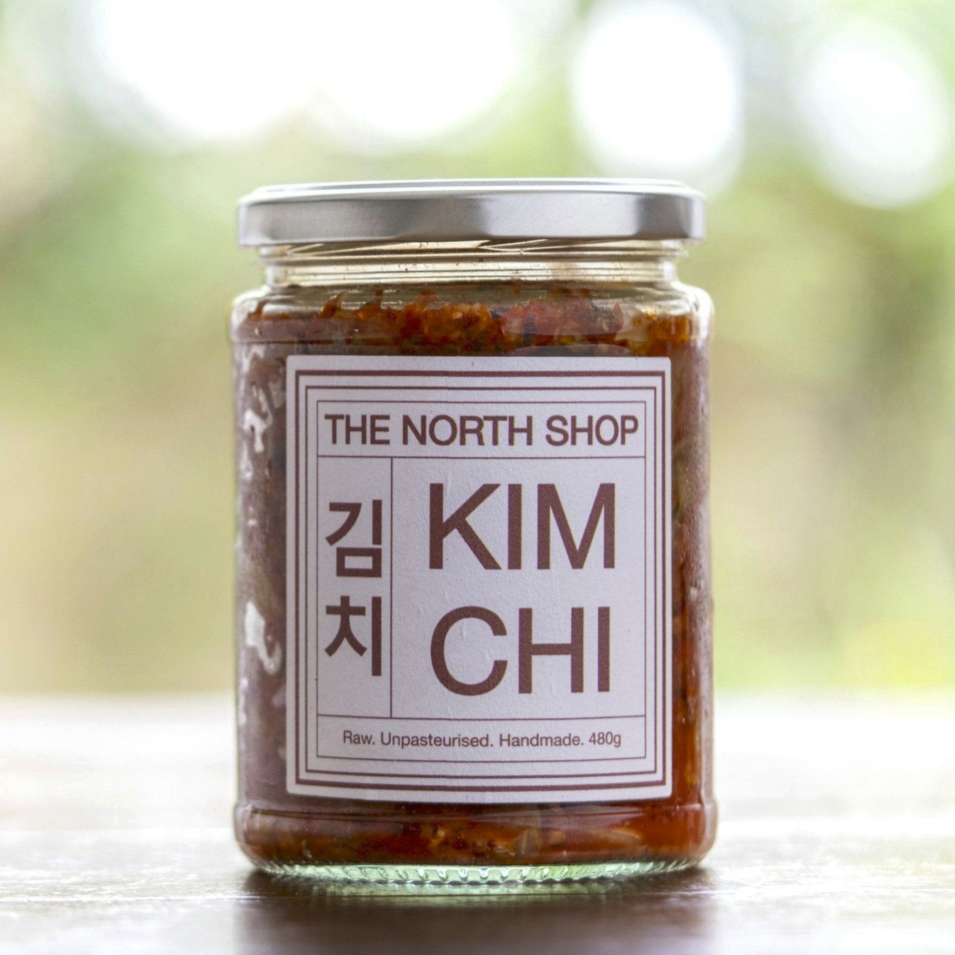 A jar of The North Shop kimchi on a wooden table with the soft image of a garden in the background.