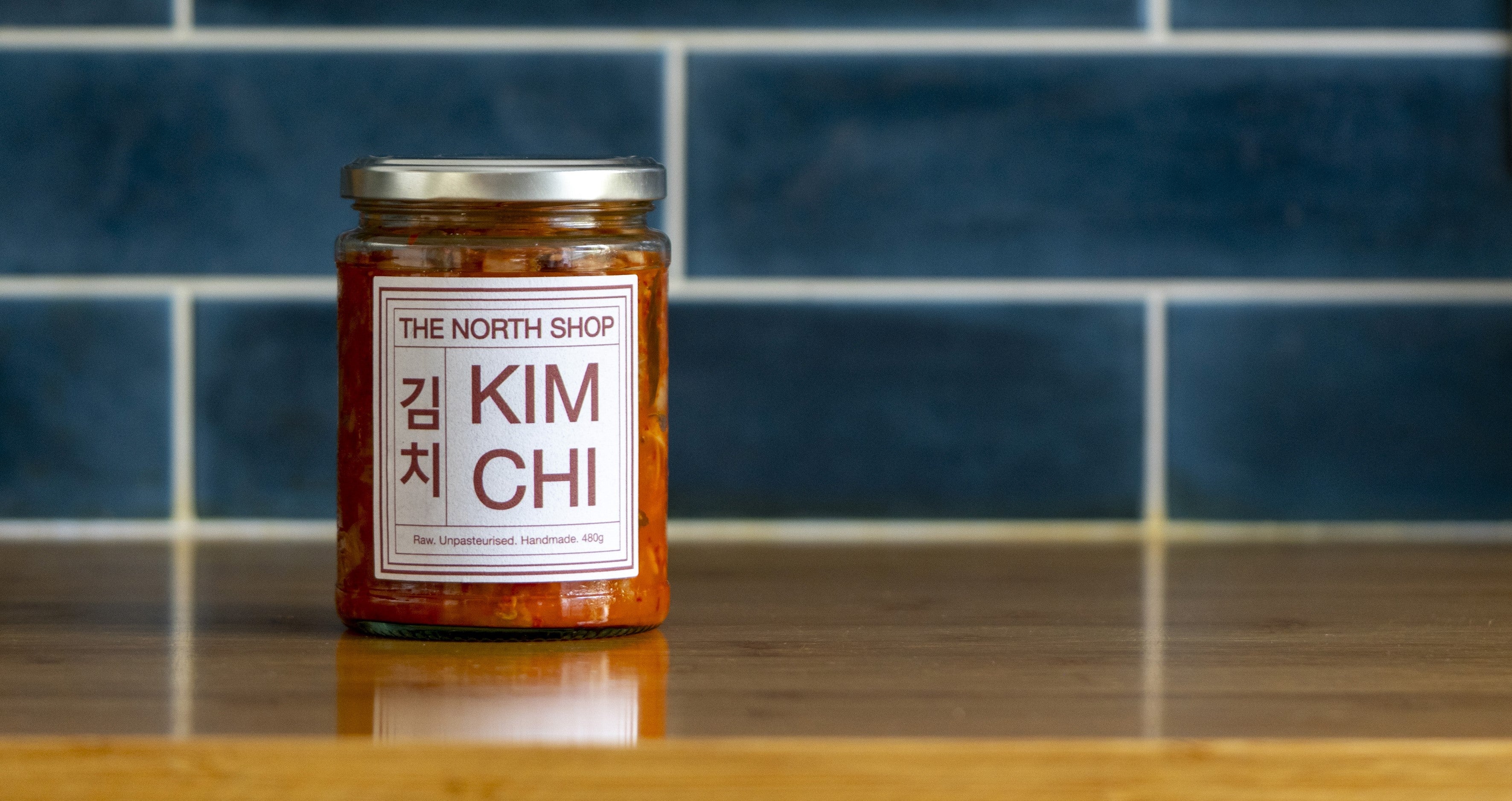 A jar of The North Shop Kimchi on a wooden counter top in front of a blue tiled wall.