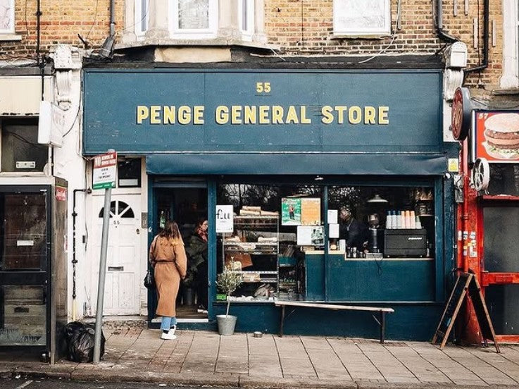 Penge General Store storefront on a street, with a dark teal awning and yellow lettering, a person standing in the doorway, and a brick building facade above.