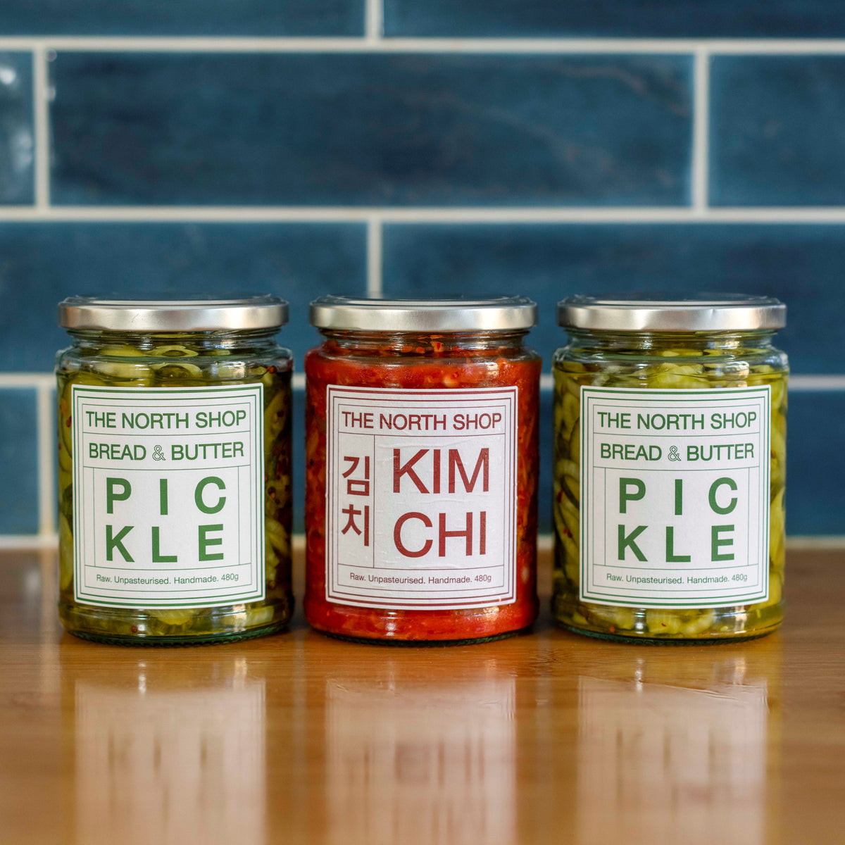 Three glass jars of The North Shop Pickles lined up on a wooden surface against a blue tiled background. The left and right jars contain green bread and butter pickles with white labels, and the center jar contains red Kimchi with a white and red label.