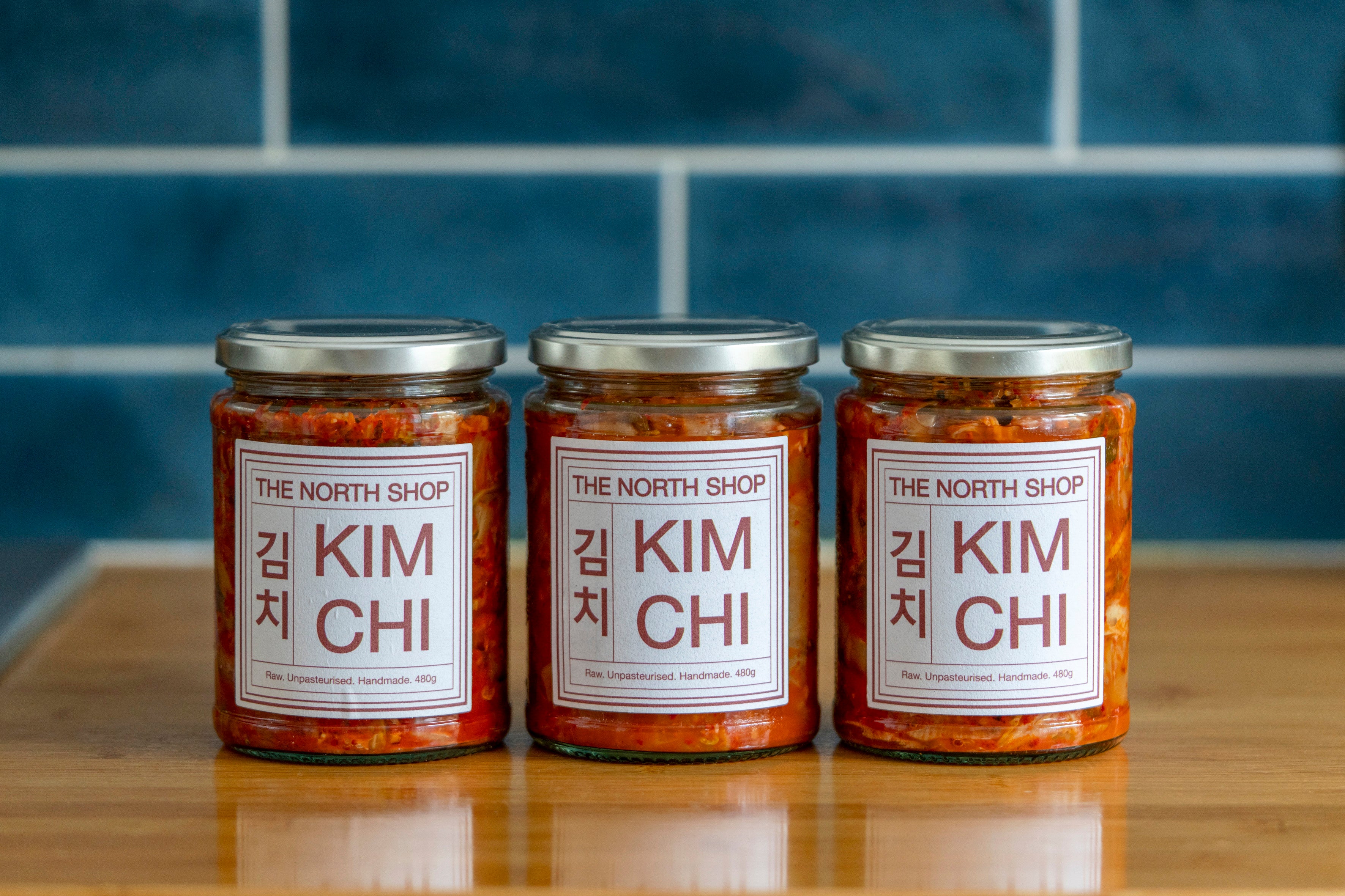 Three jars of The North Shop kimchi on a wooden work surface in front of a blue tiled wall.