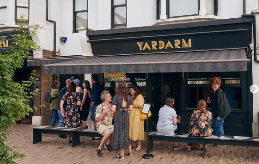 A real outdoor storefront scene with several people gathered and sitting on benches in front of a shop labeled YARDARM. Because people and a real environment are clearly present, this is Lifestyle rather than Product/Packaging.
